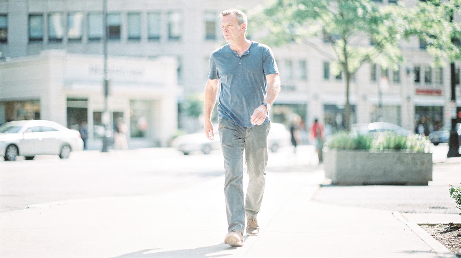 A man walks on a sunny city sidewalk with buildings and parked cars in the background.