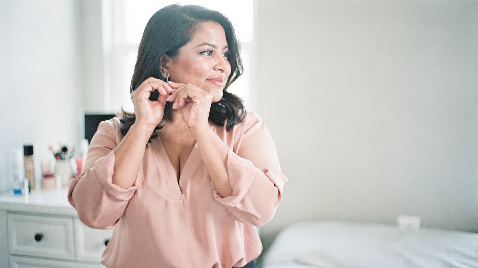 A woman in a pink blouse stands in a bedroom putting on earrings.