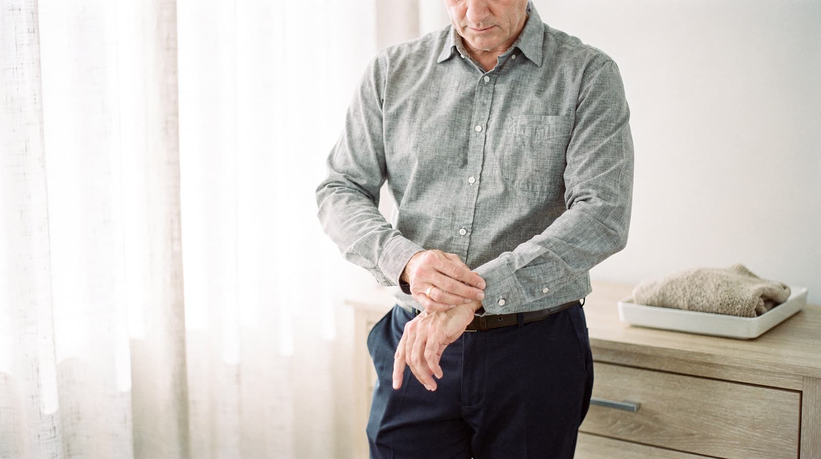 A man in a gray shirt adjusts his sleeve while standing in a bright room with a dresser and folded clothes.