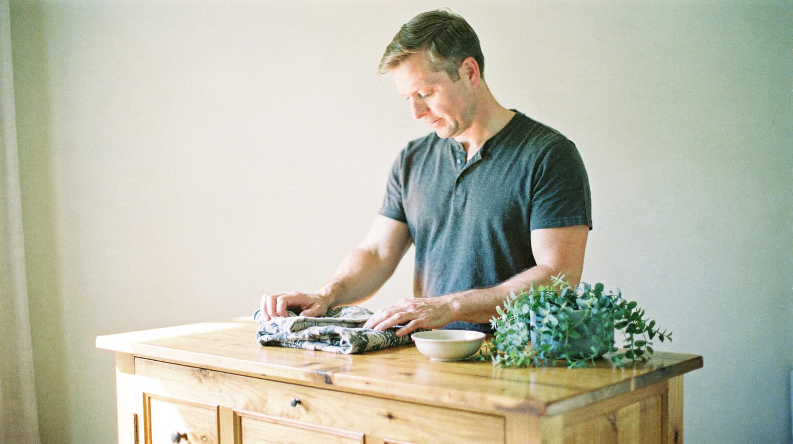 A man folds clothes on a wooden table next to a small plant and a bowl.