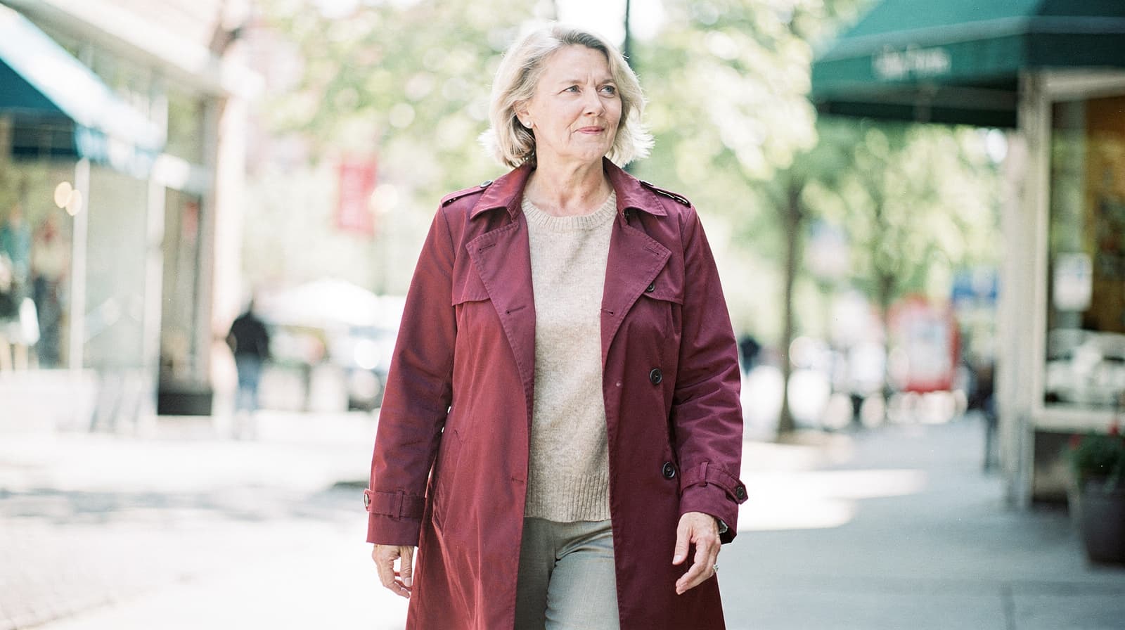 A woman in a red coat walks down a sunny city street lined with shops and trees.