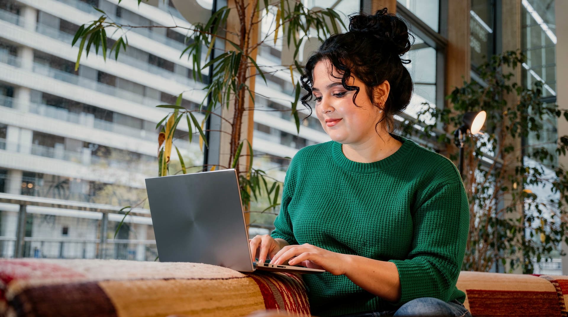 A woman in a green sweater types on a laptop while sitting on a patterned couch in a modern building with large windows.