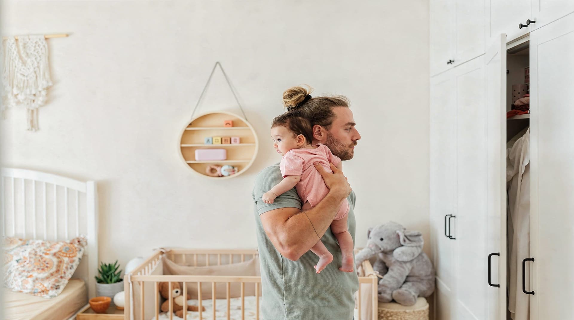 A man holds a baby in a nursery with a crib, toys, and a wall shelf in the background.