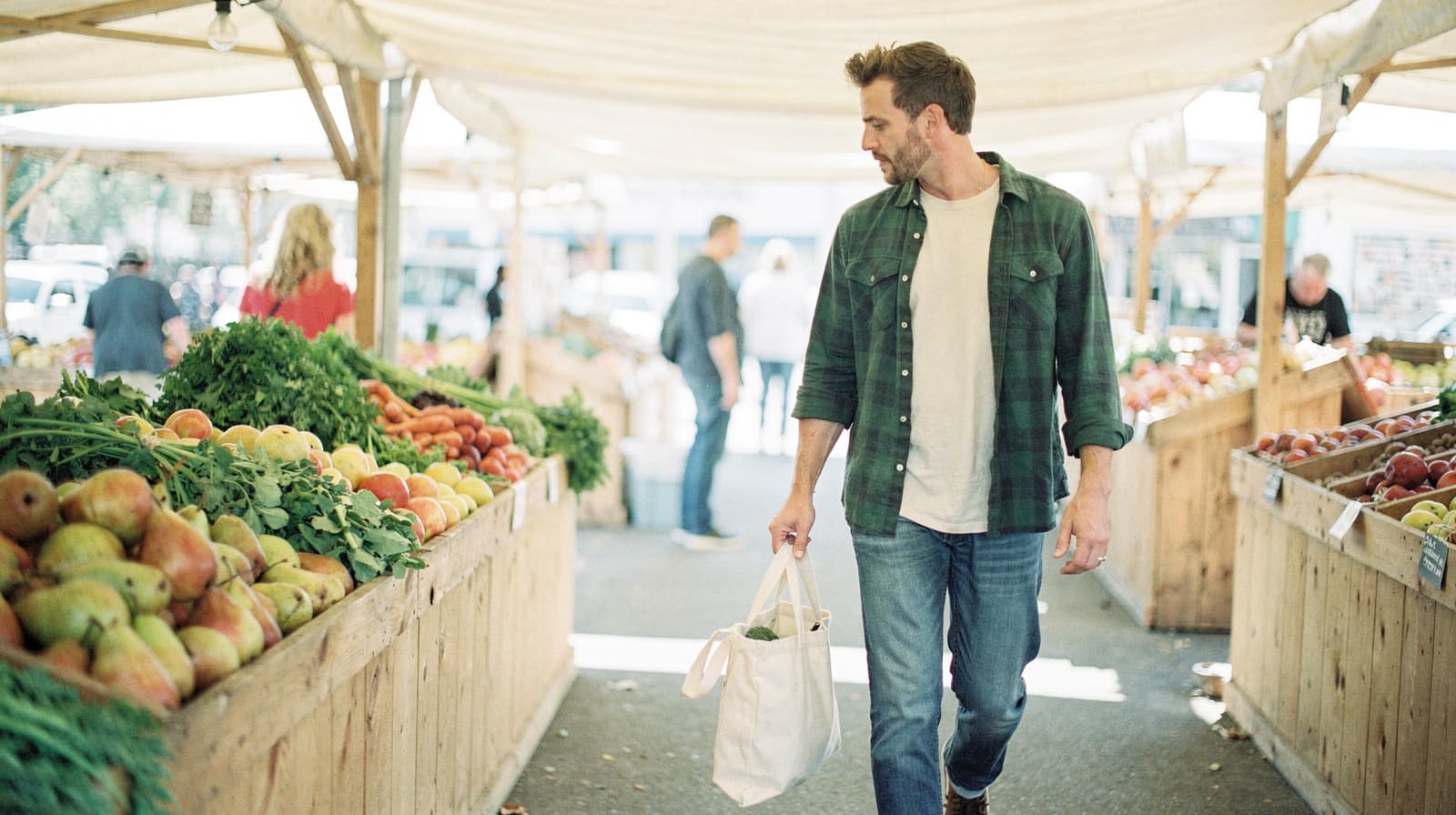 A man carrying a tote bag walks through an outdoor market with wooden stalls filled with fruits and vegetables.