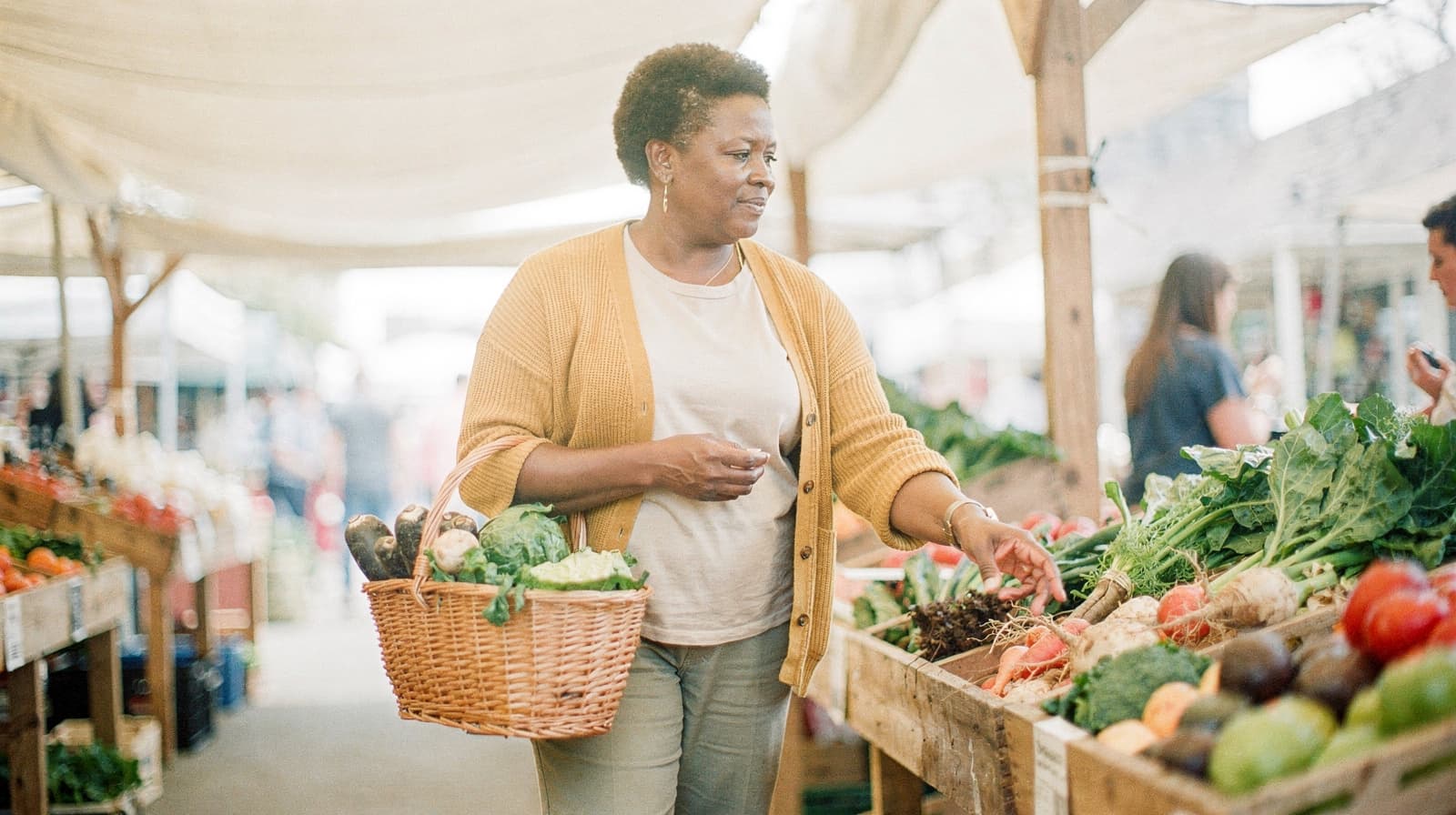 A woman in a yellow cardigan selects vegetables from a market stall while holding a wicker basket.