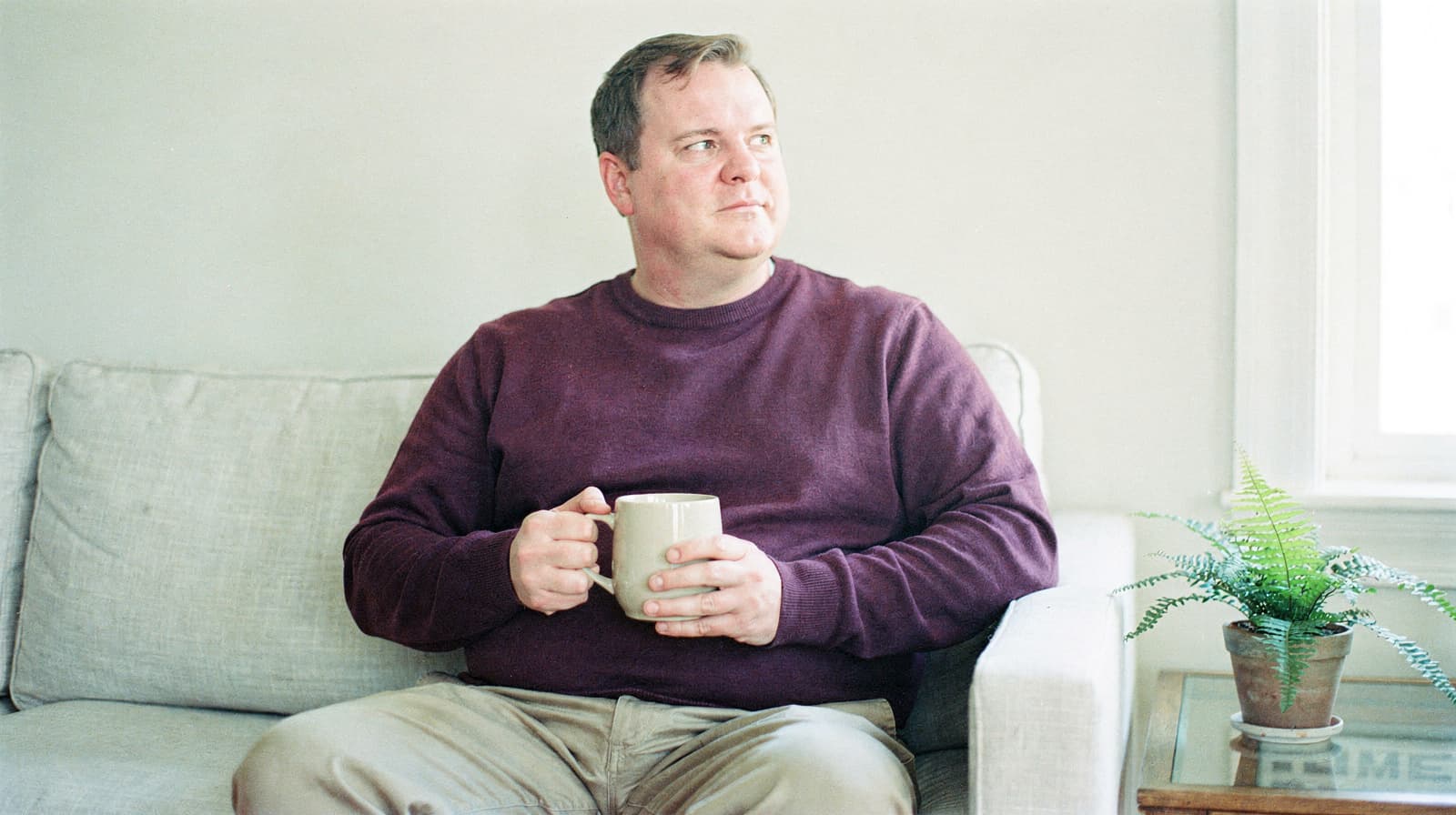 A man sits on a couch holding a mug, looking to the side, with a small plant on a table nearby.