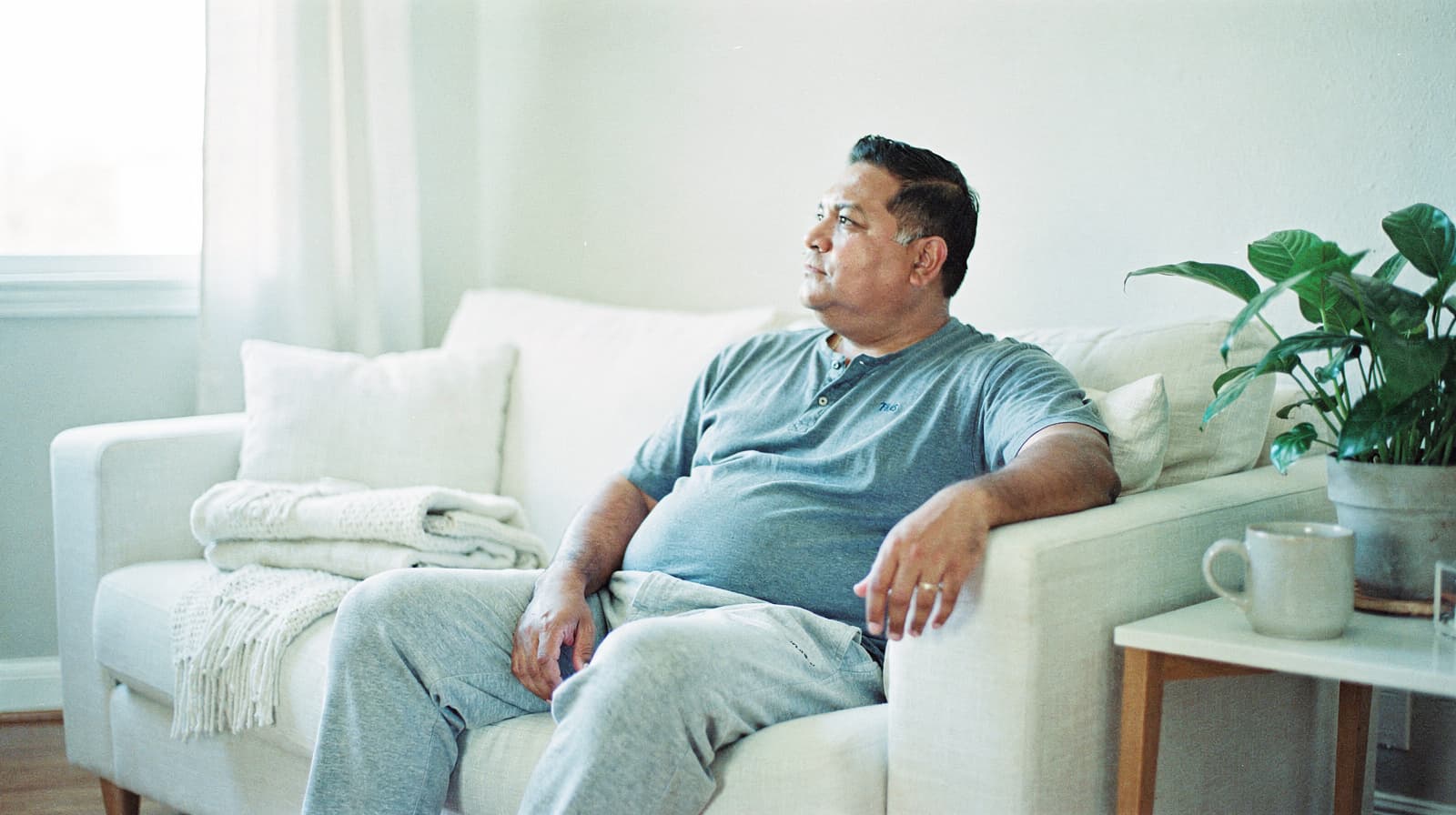 A man sits on a sofa in a living room, looking out the window with a plant and a mug on a nearby table.
