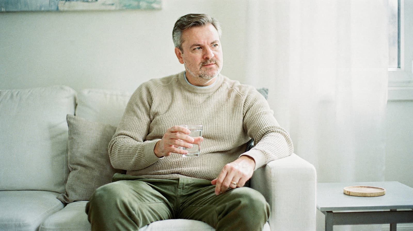 A man sits on a couch holding a glass of water, looking thoughtfully out a window.