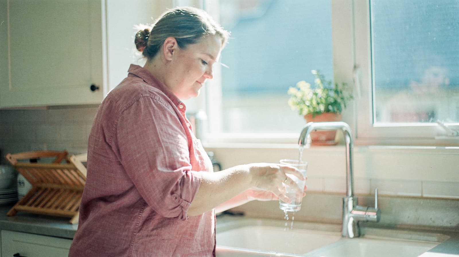 A woman in a pink shirt washes a glass at a kitchen sink with sunlight streaming through the window.