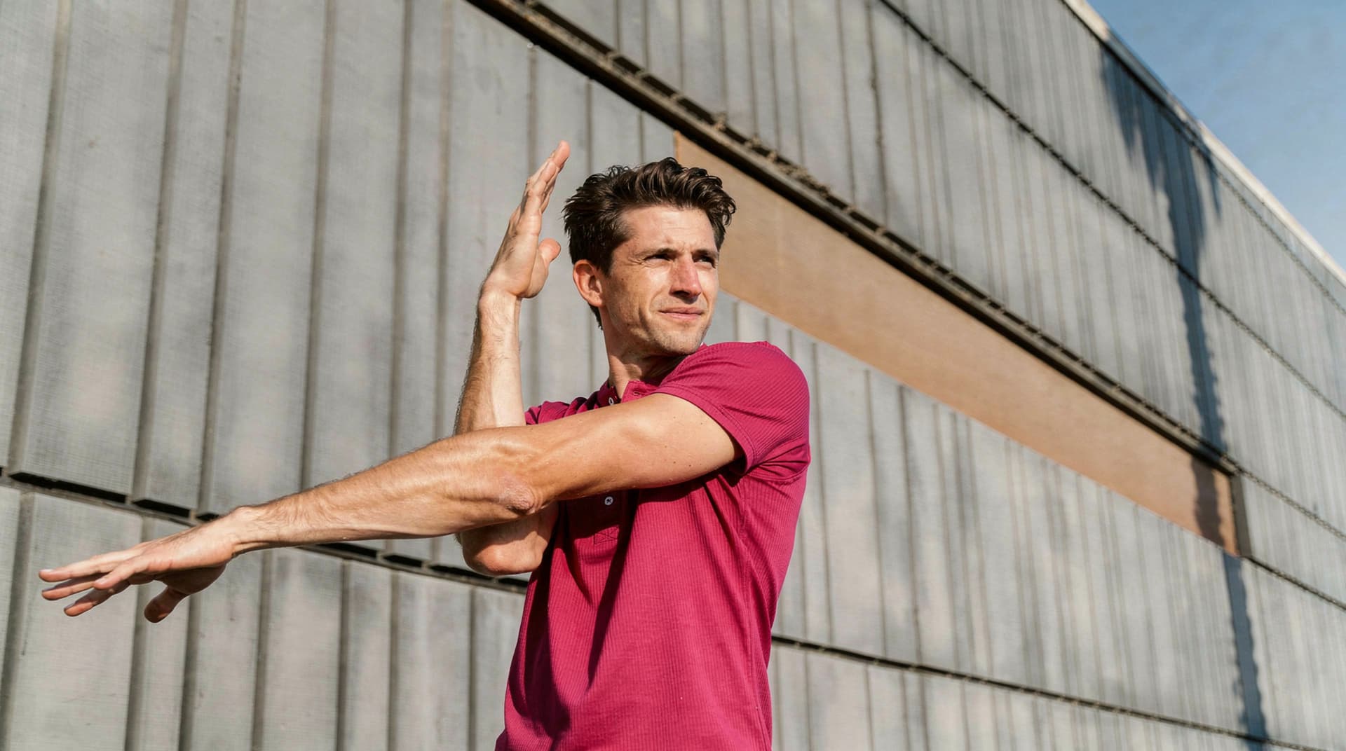 A man in a pink shirt stretches his arms in front of a large metal wall under a clear blue sky.