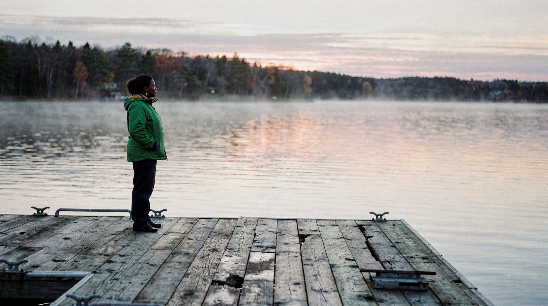 A person in a green jacket stands on a wooden dock overlooking a misty lake at sunrise.