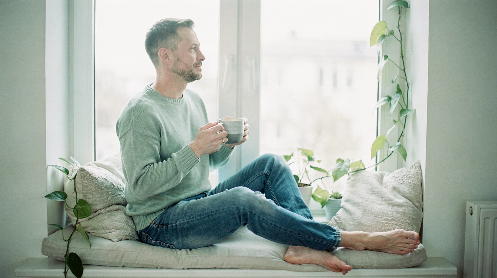 A man sits on a windowsill holding a mug, surrounded by cushions and plants.