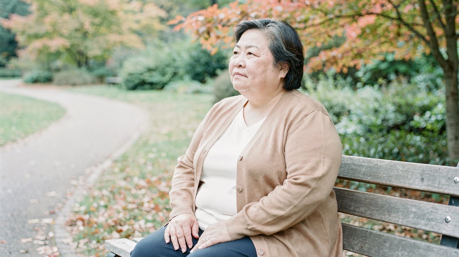 A woman sits on a park bench surrounded by autumn trees and fallen leaves.