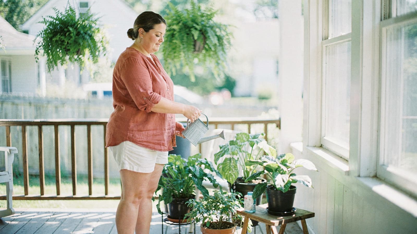 A woman waters potted plants on a sunlit porch with hanging ferns in the background.