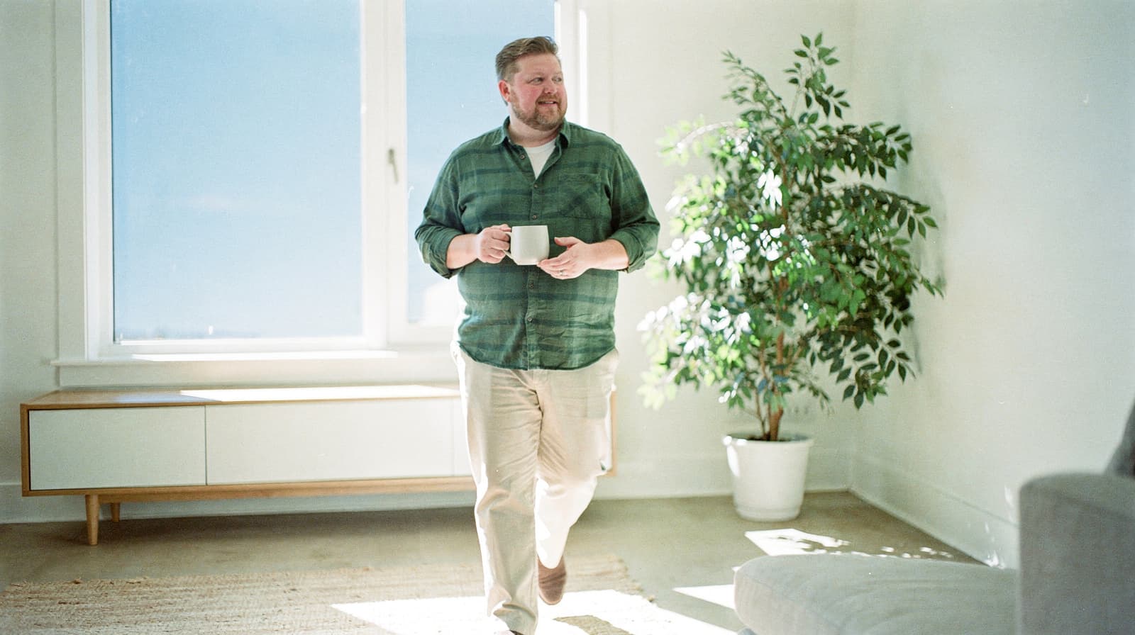 A man holding a mug walks through a sunlit room with a large window and a potted plant.