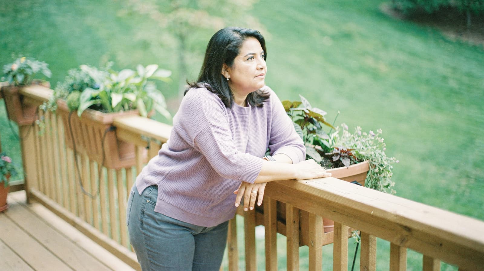 A woman in a purple sweater leans on a wooden deck railing, surrounded by potted plants, looking out at a grassy yard.