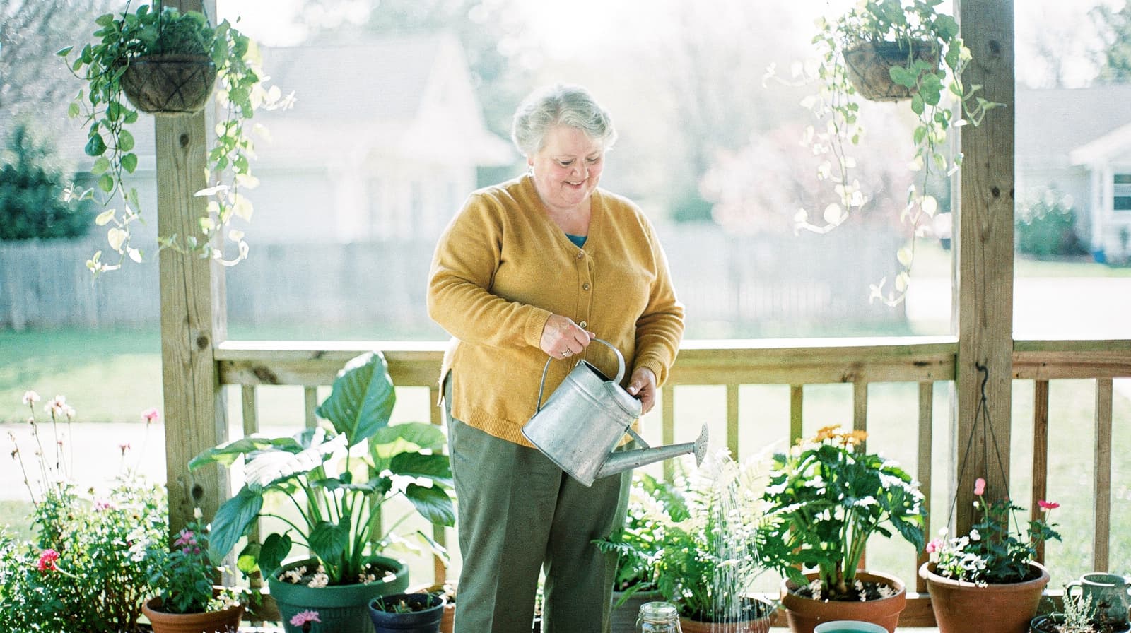 A woman waters potted plants on a wooden deck surrounded by hanging and potted greenery.