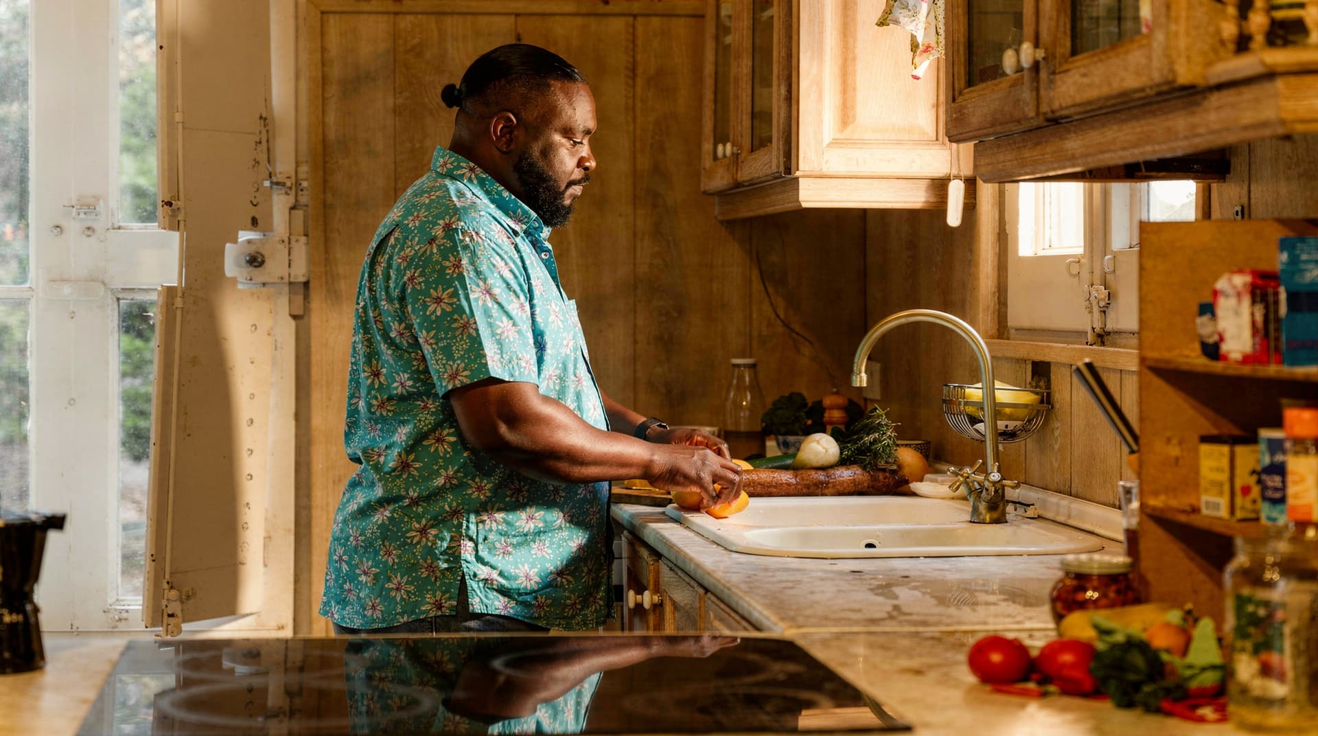A man in a floral shirt washes vegetables at a kitchen sink with sunlight streaming through the window.