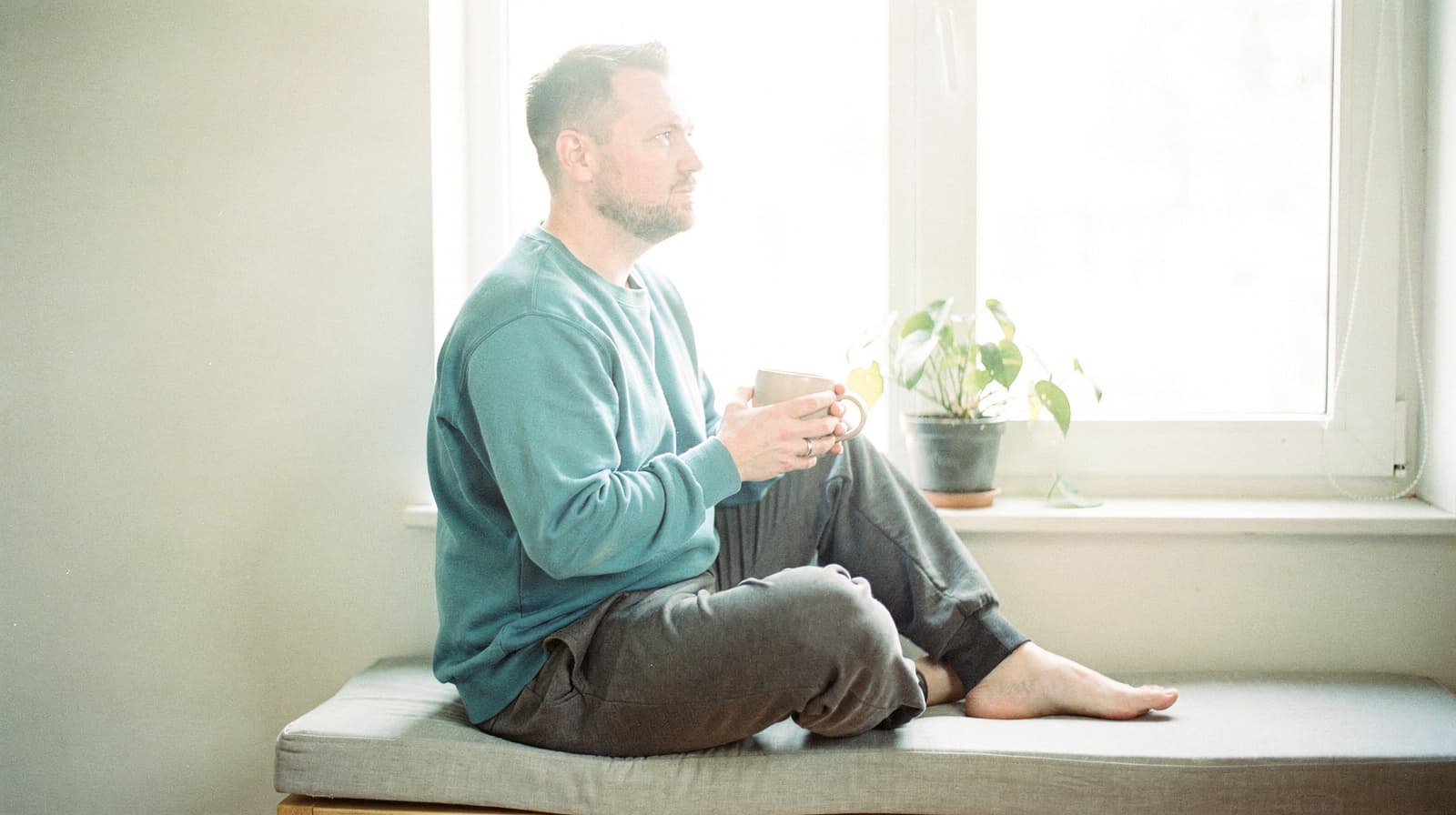 A man sits on a cushioned bench by a window holding a mug, with a potted plant nearby.
