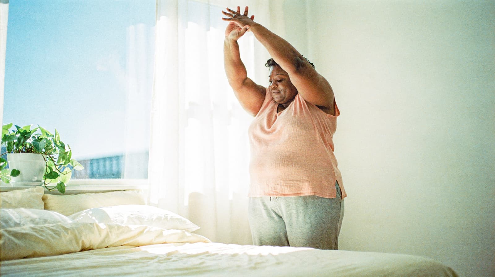 A person stretches with arms raised beside a bed in a sunlit room.