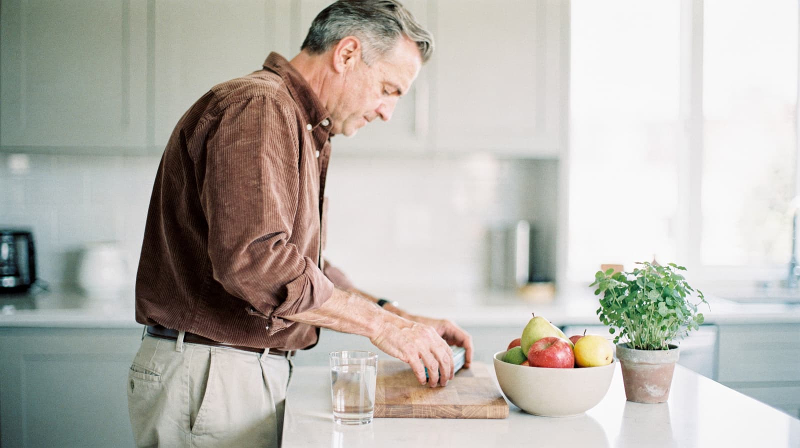 A man slices a vegetable on a wooden cutting board in a bright kitchen with a bowl of fruit and a potted plant nearby.
