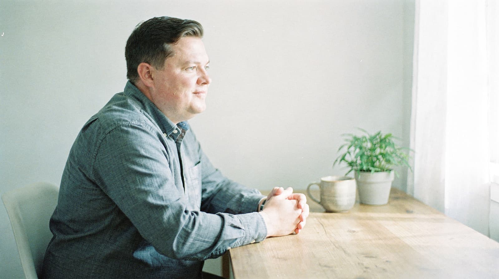 A man sits at a wooden table with a plant and a mug, looking out a window.