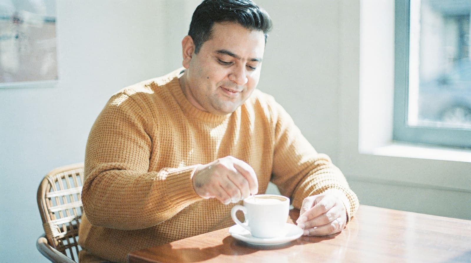 A man in a sweater stirs a cup of coffee at a wooden table near a window.