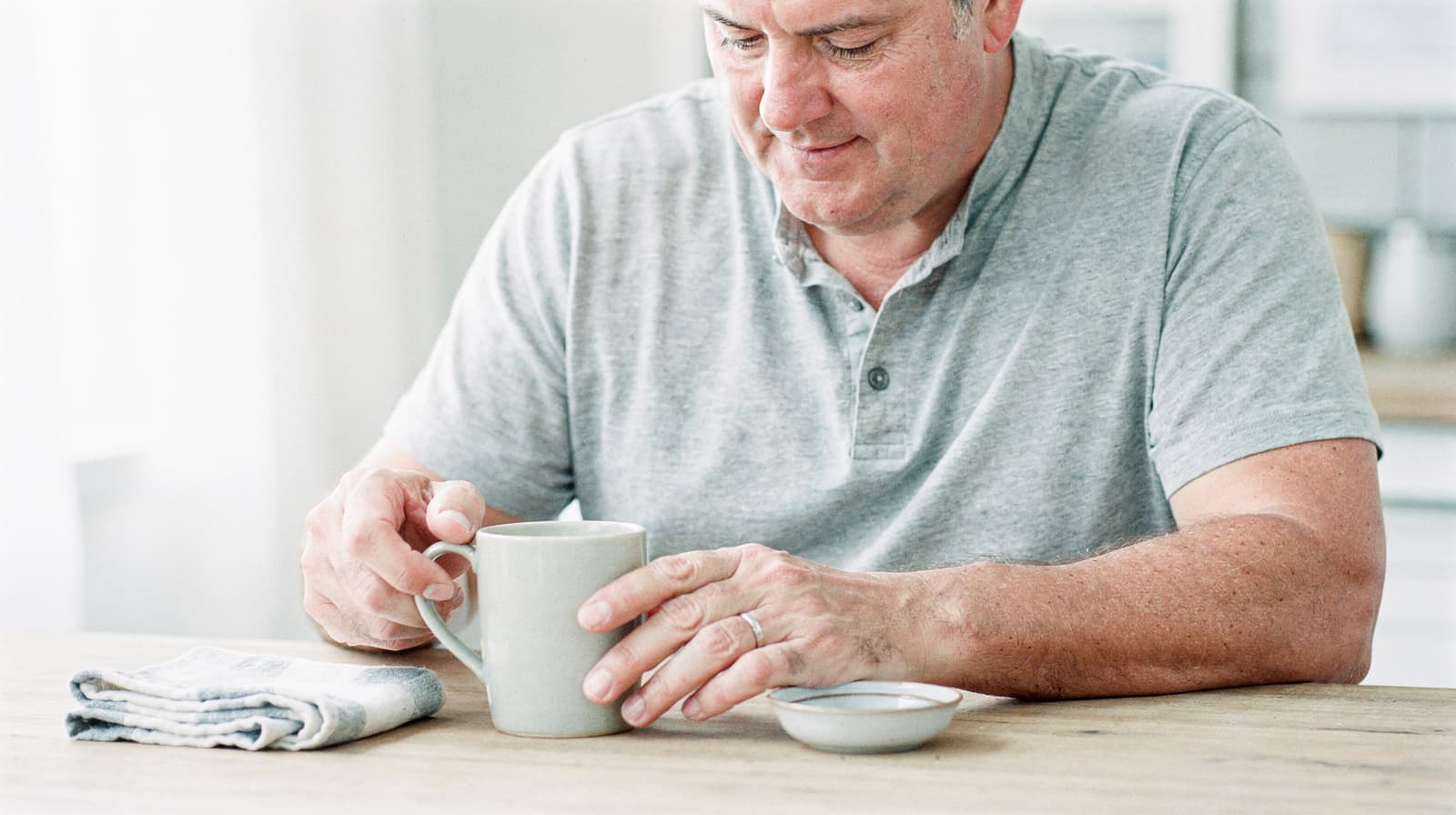 A man in a gray shirt sits at a wooden table holding a mug with a small dish and a folded cloth nearby.
