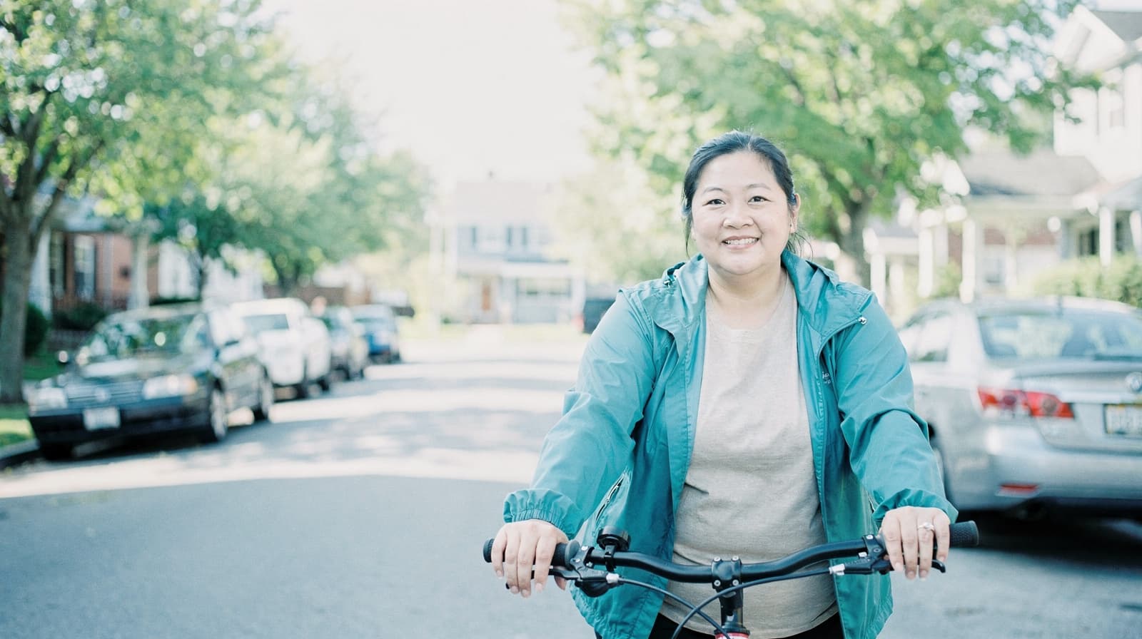 A woman in a teal jacket rides a bicycle on a suburban street lined with parked cars and trees.