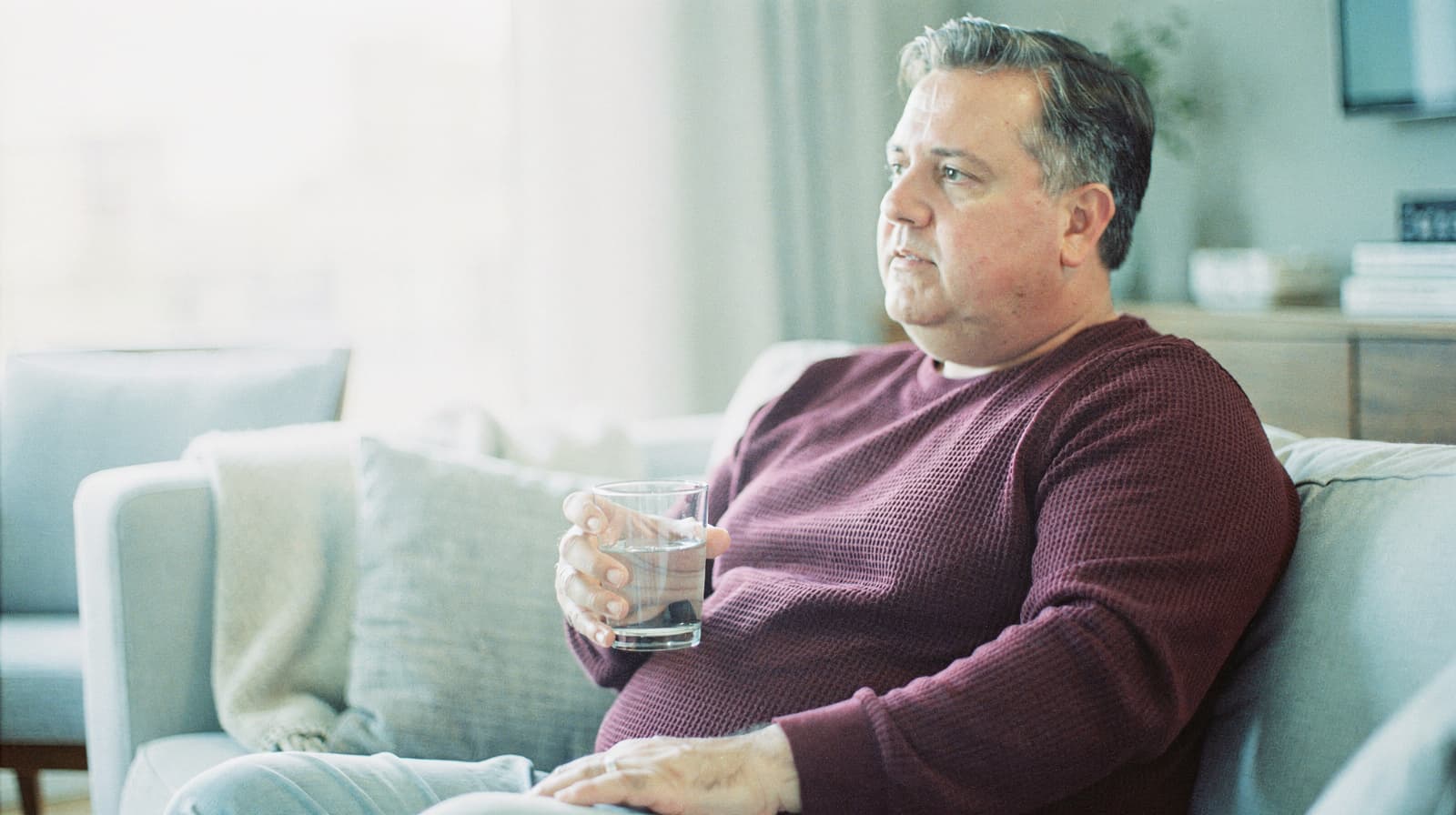 A man sits on a couch holding a glass of water in a softly lit living room.