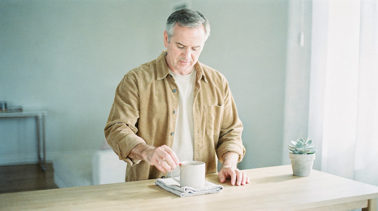A man stirs a drink in a mug on a wooden table next to a small potted plant in a bright room.