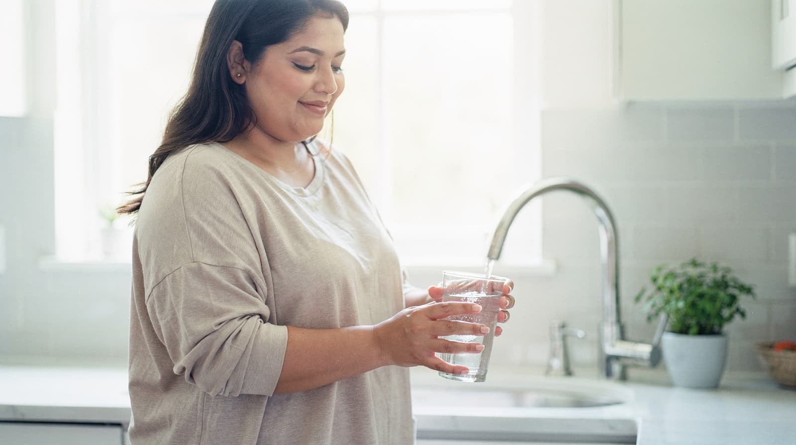 A woman stands in a kitchen holding a glass of water near a sink.