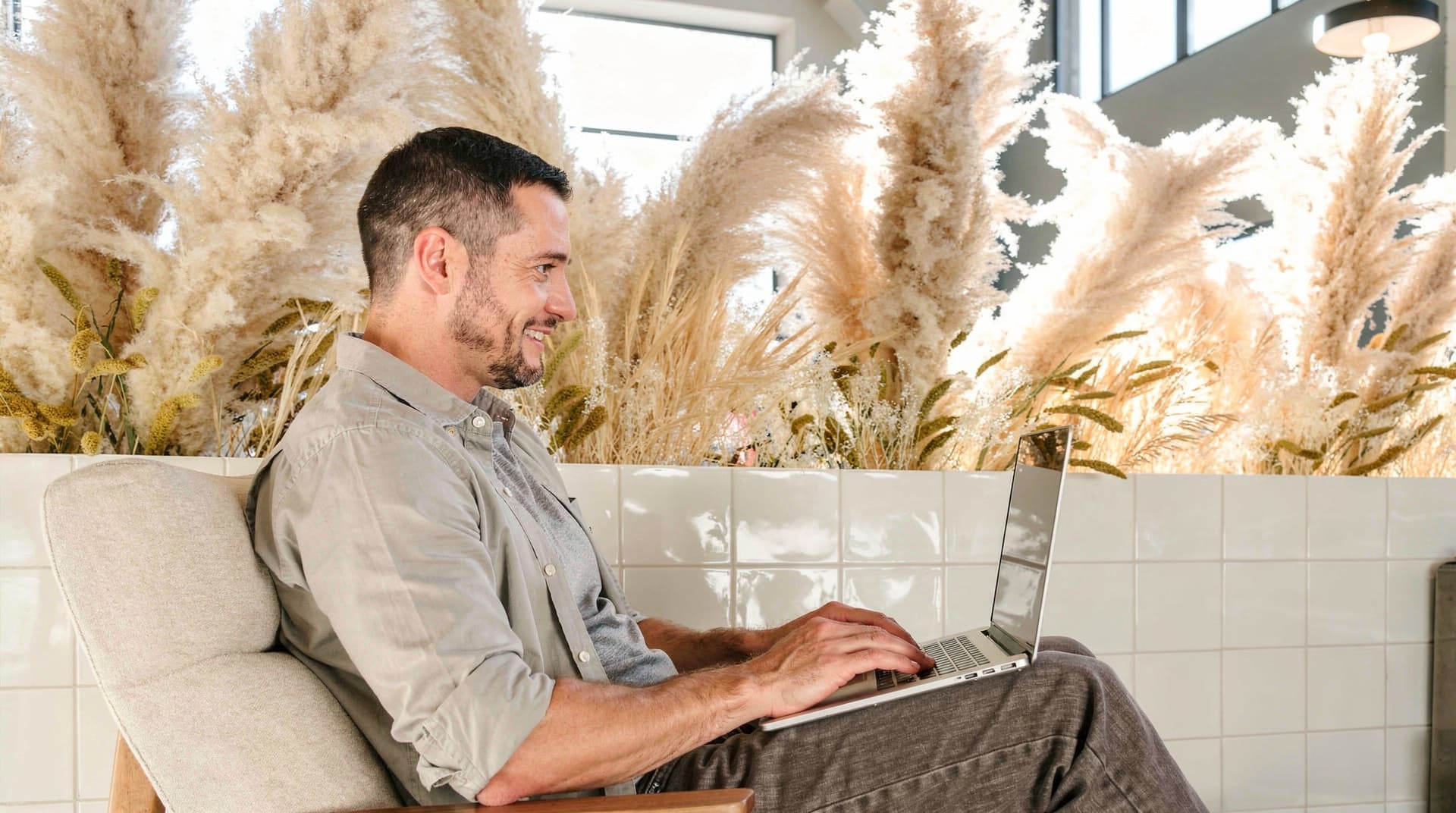 A man sits in a chair using a laptop, surrounded by tall pampas grass indoors.