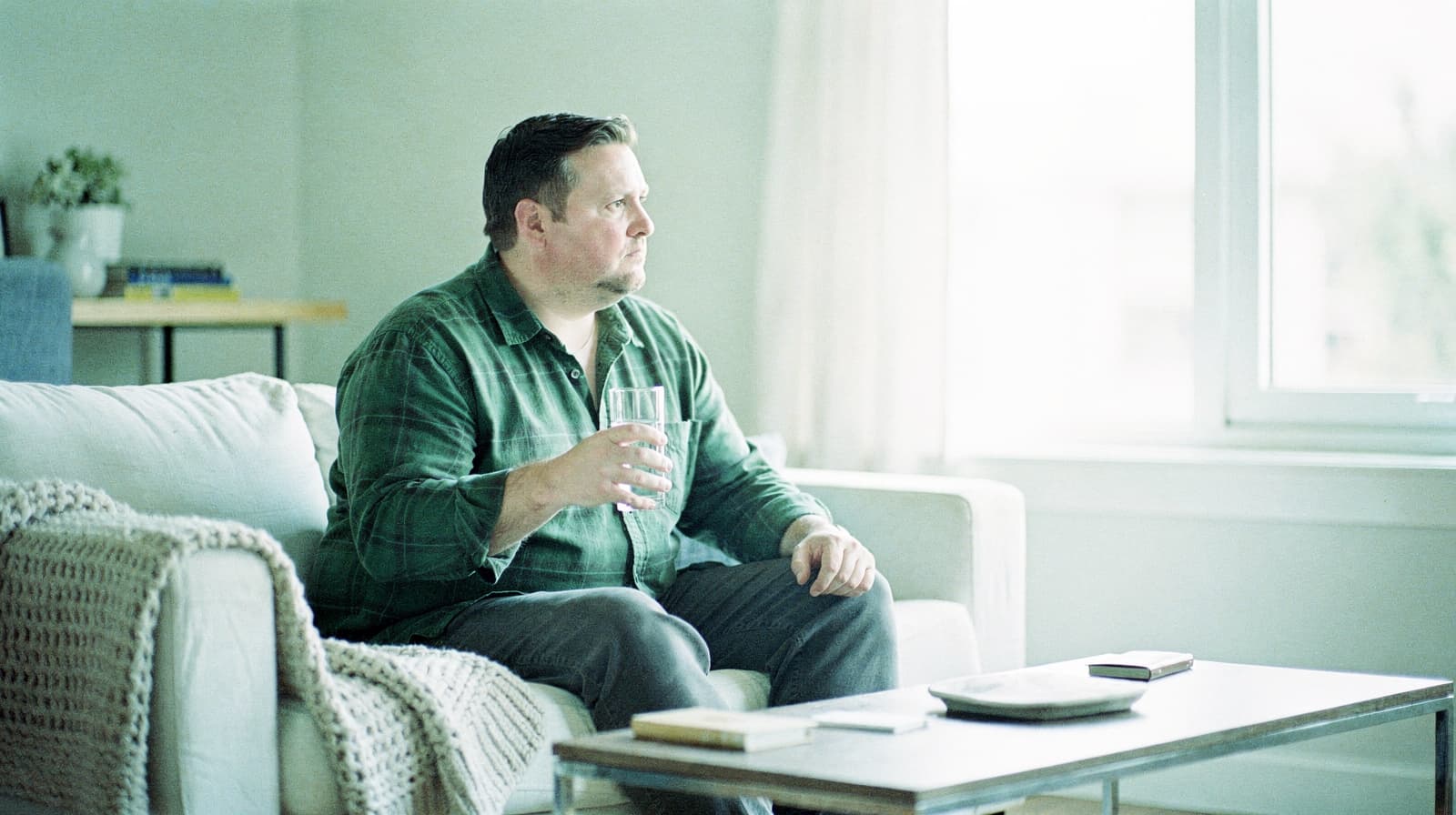 A man in a green shirt sits on a couch holding a glass of water, looking out a window.