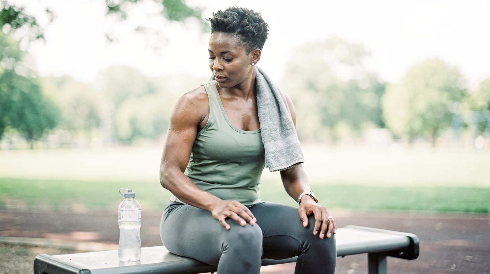A woman sits on a bench outdoors with a towel around her neck and a water bottle beside her.