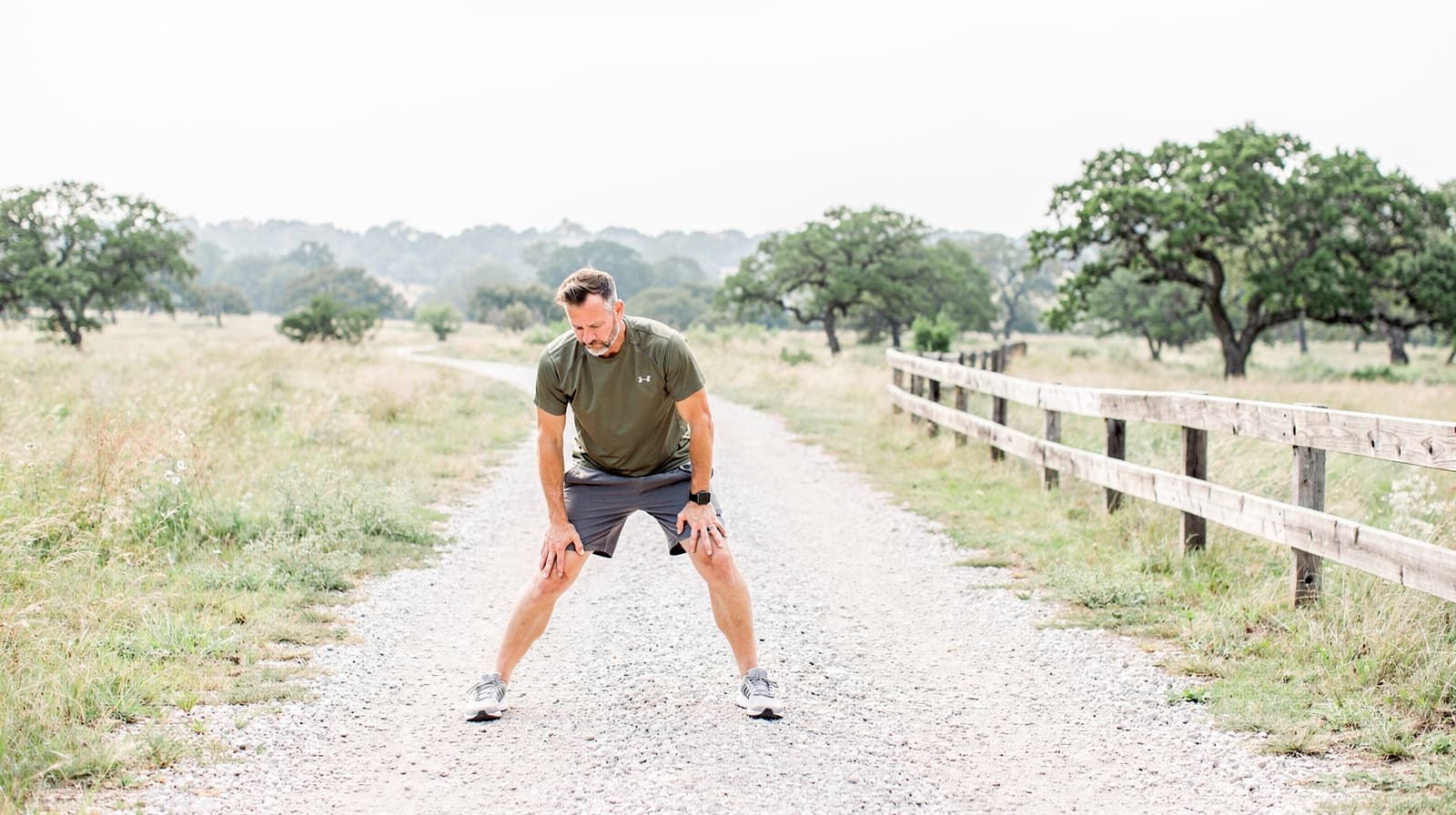 A man stretches on a gravel path in a grassy field with scattered trees and a wooden fence.