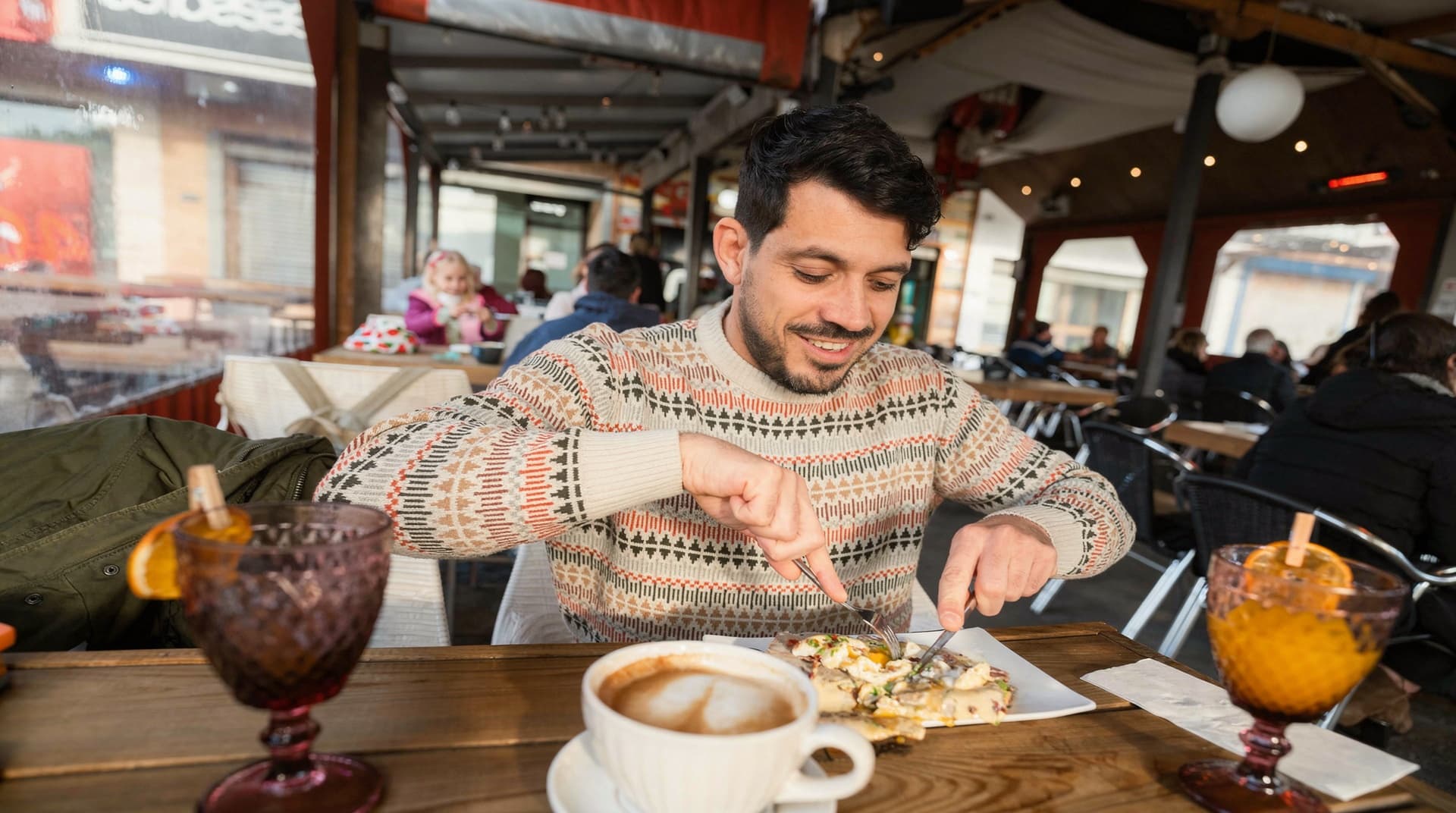 A man in a patterned sweater eats a meal at an outdoor café with a coffee and two drinks on the table.