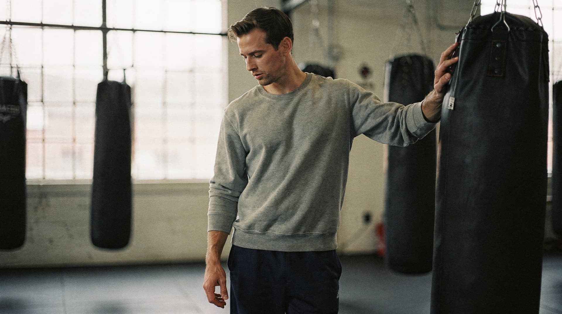 A man in a gray sweatshirt stands beside a punching bag in a gym with large windows.