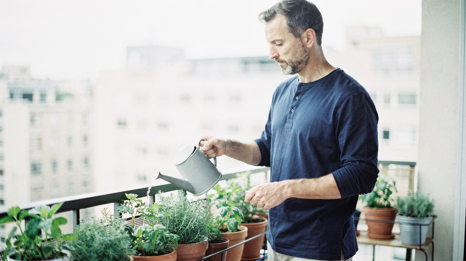 A man waters potted plants on a balcony overlooking a cityscape.