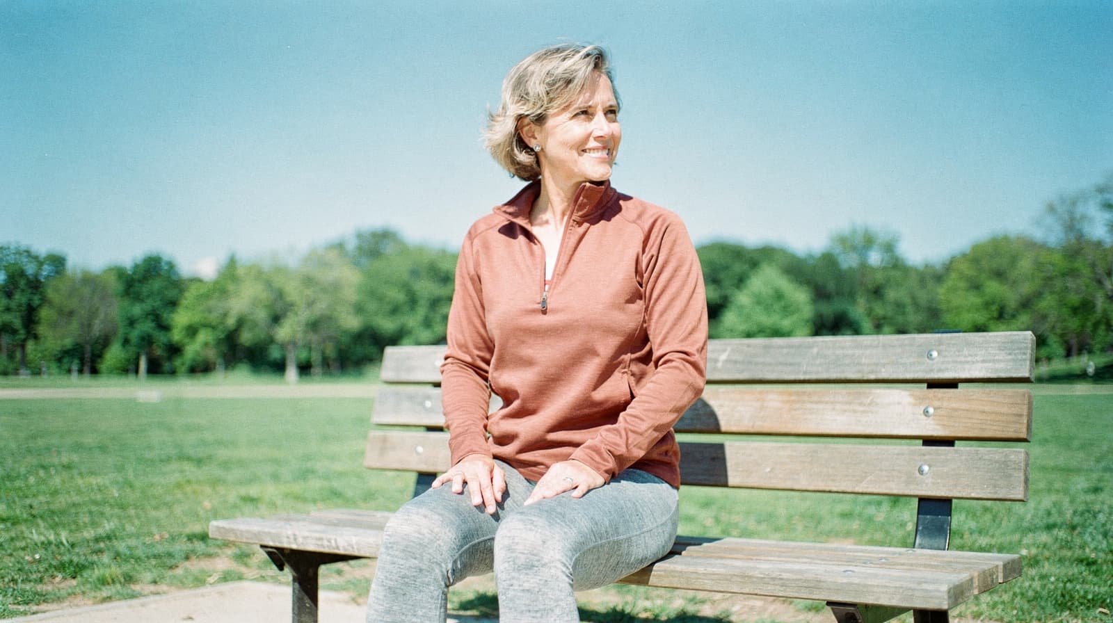 A woman sits on a wooden bench in a park, smiling and looking to the side.