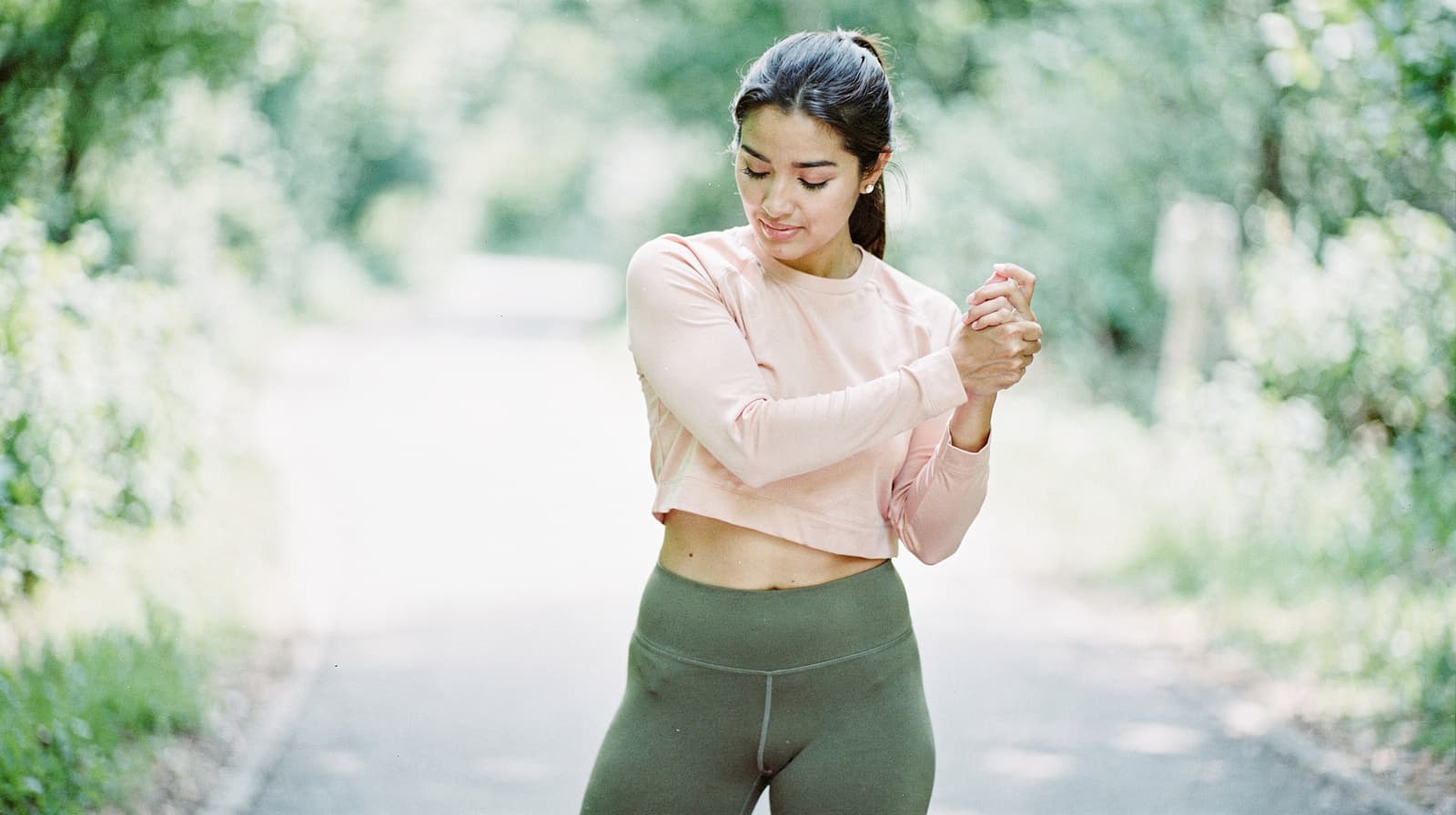 A woman in athletic wear stretches her arms on a tree-lined path.