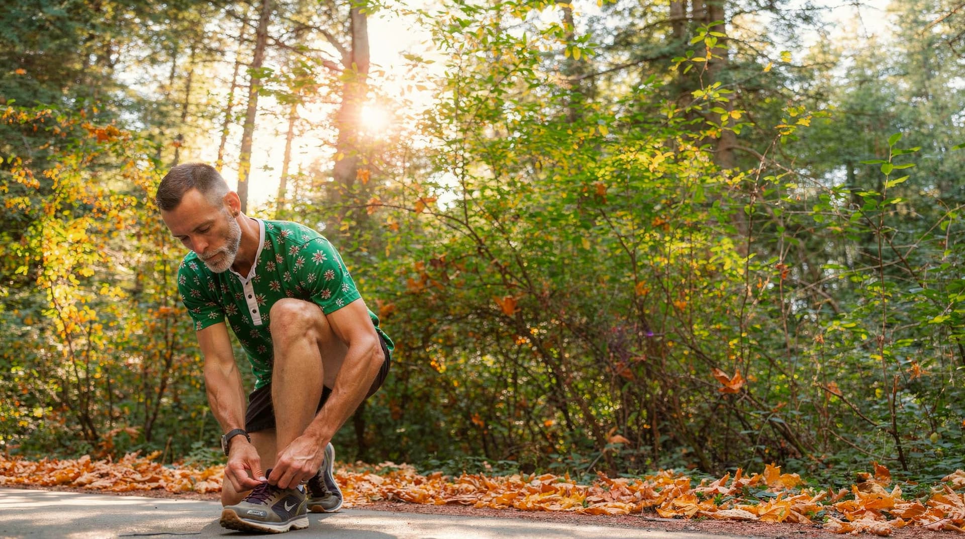 A man ties his shoe on a forest path surrounded by autumn leaves and sunlight filtering through the trees.