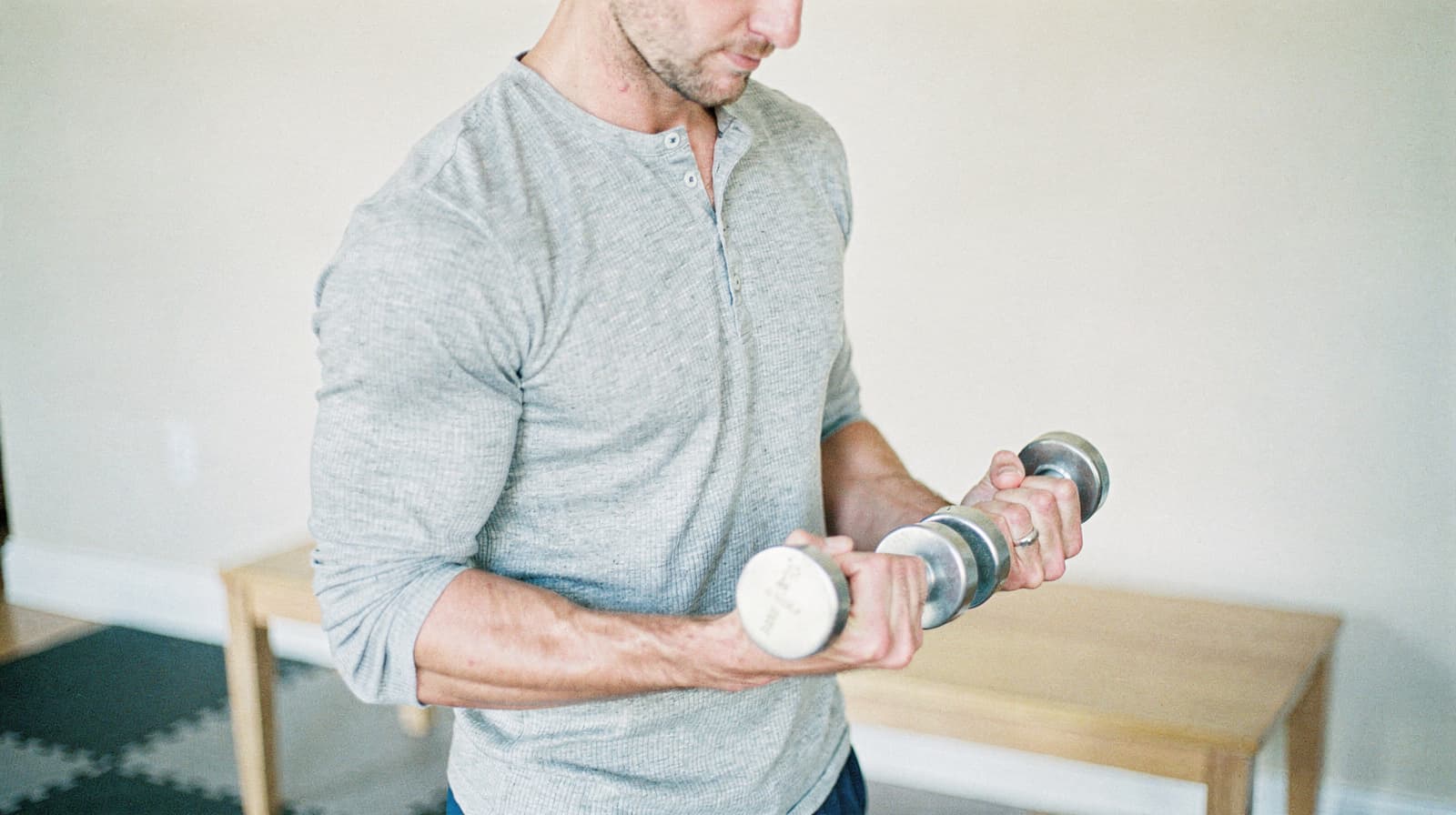 A man in a gray shirt lifts a dumbbell in a room with a wooden bench.