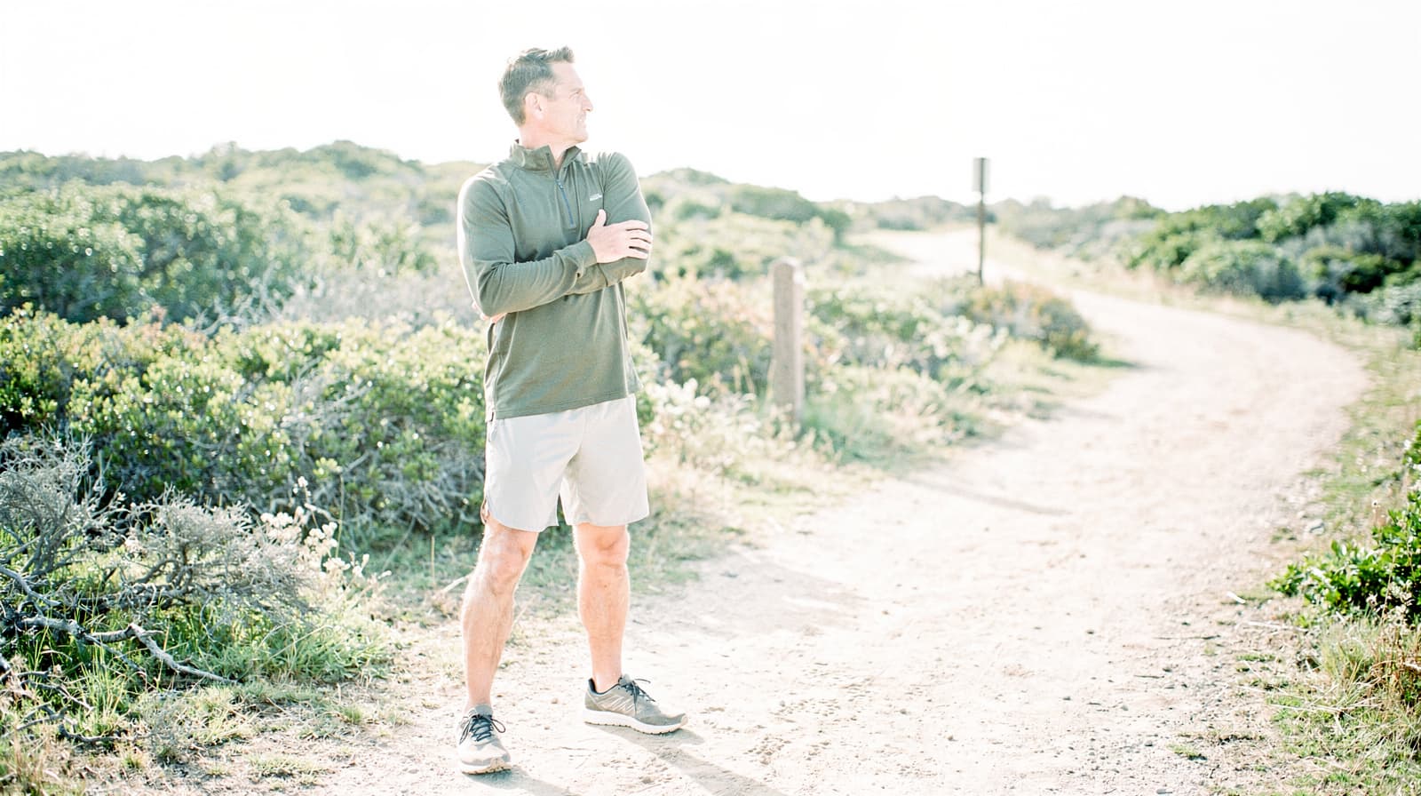 A man in athletic wear stretches his arm while standing on a dirt path surrounded by greenery.