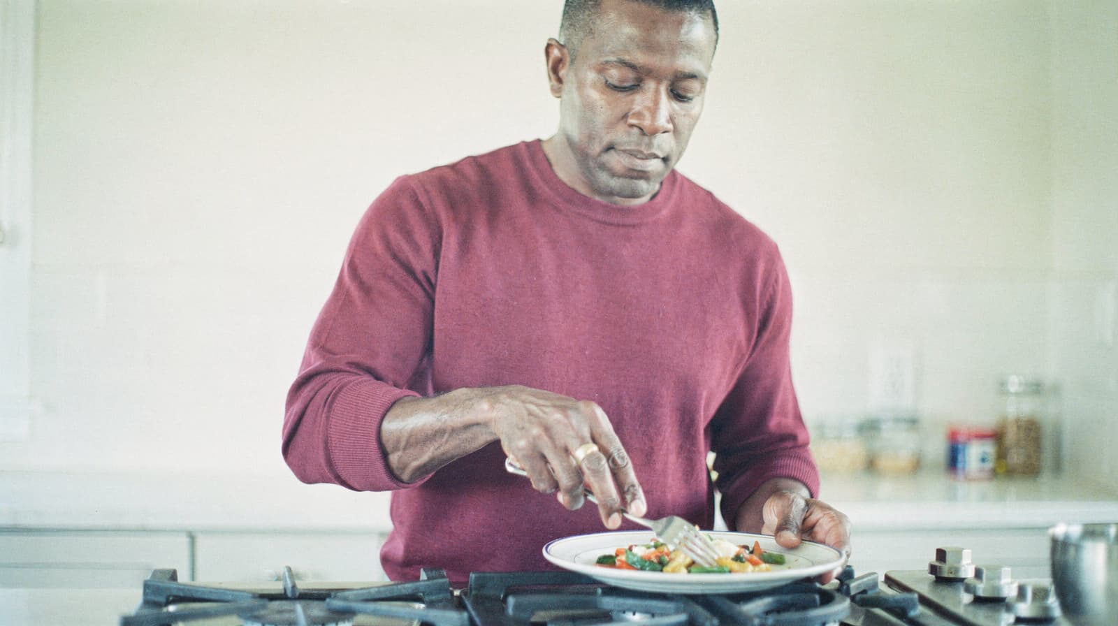 A man in a red sweater prepares a meal on a plate in a kitchen.