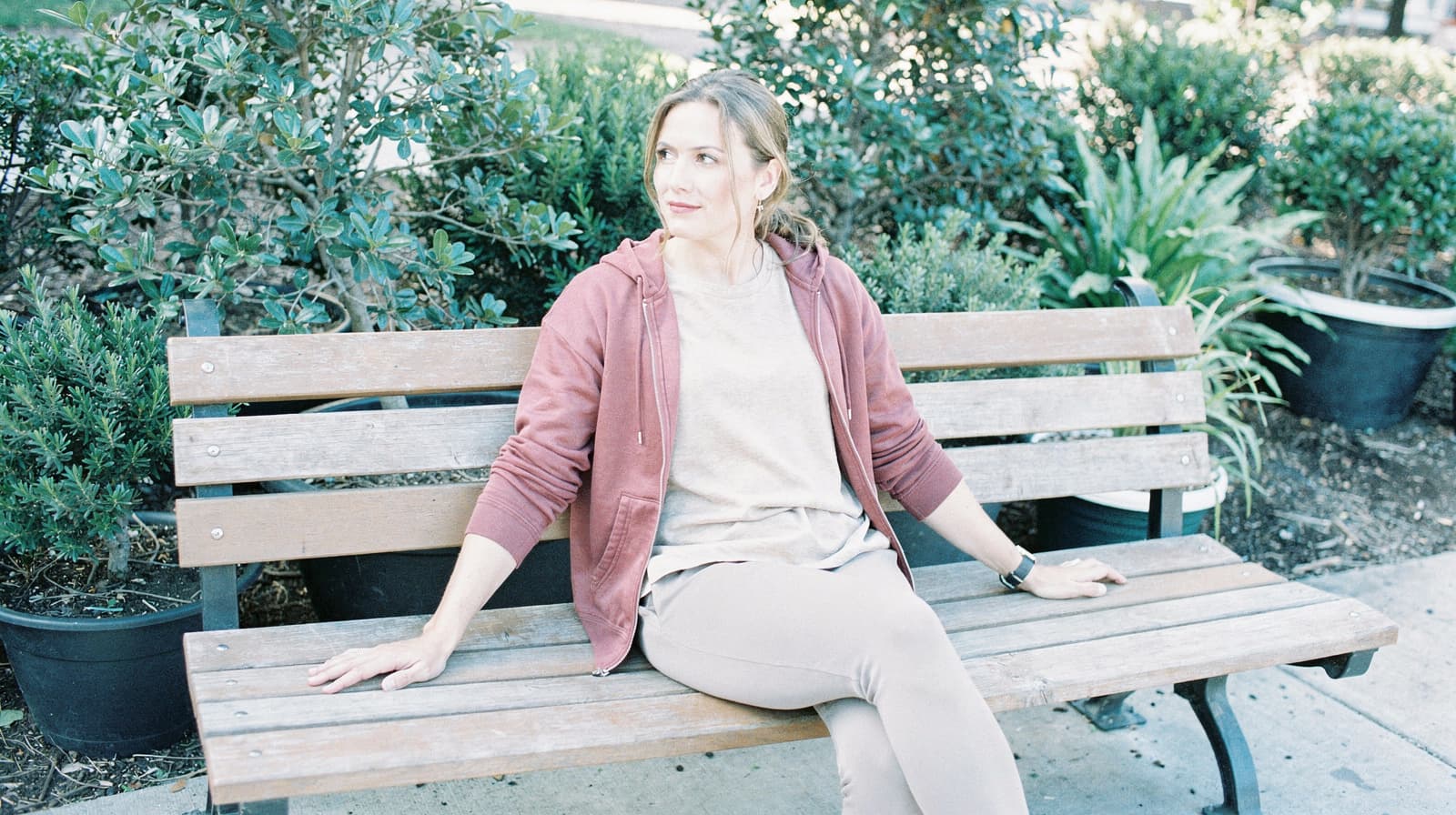 A woman sits on a wooden bench in a garden, surrounded by green plants.