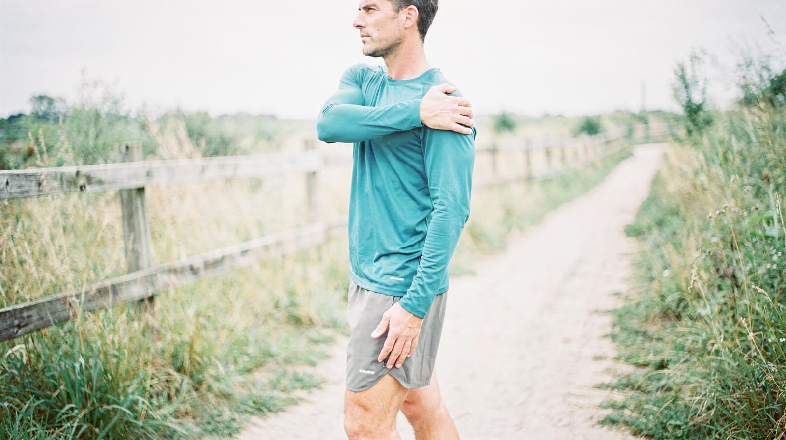 A man in a blue long-sleeve shirt stretches his arm while standing on a dirt path surrounded by grass and wooden fences.