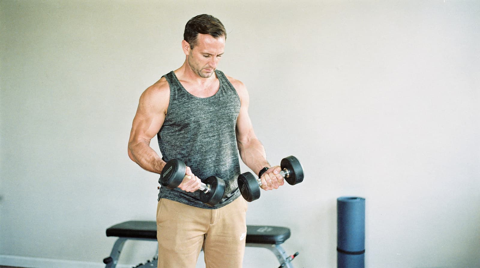 A man in a sleeveless shirt lifts dumbbells in a gym with a bench and exercise mat in the background.