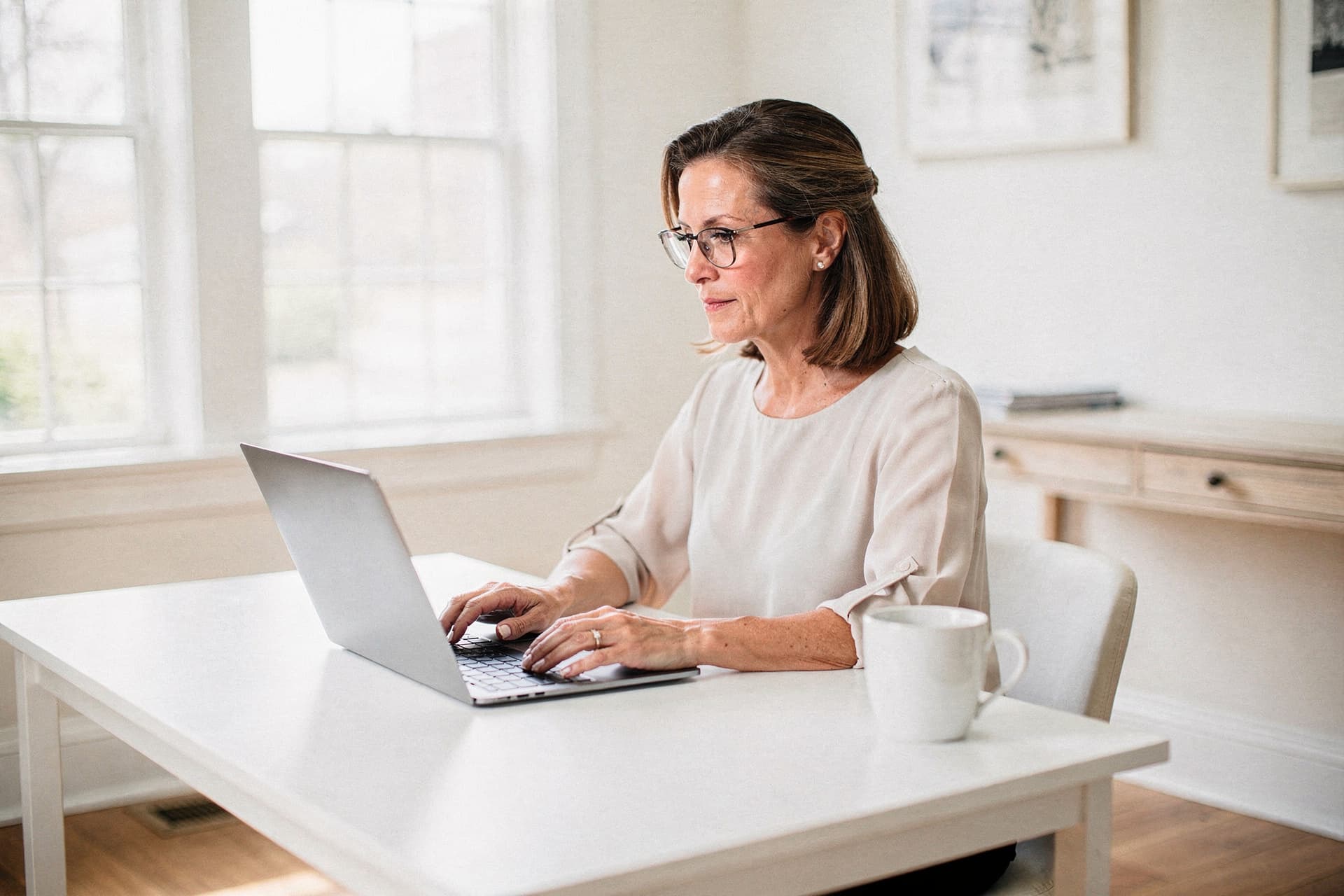 A woman with glasses reads on a laptop at a white desk beside tall windows in a bright, sunlit home office, a white mug at her side.