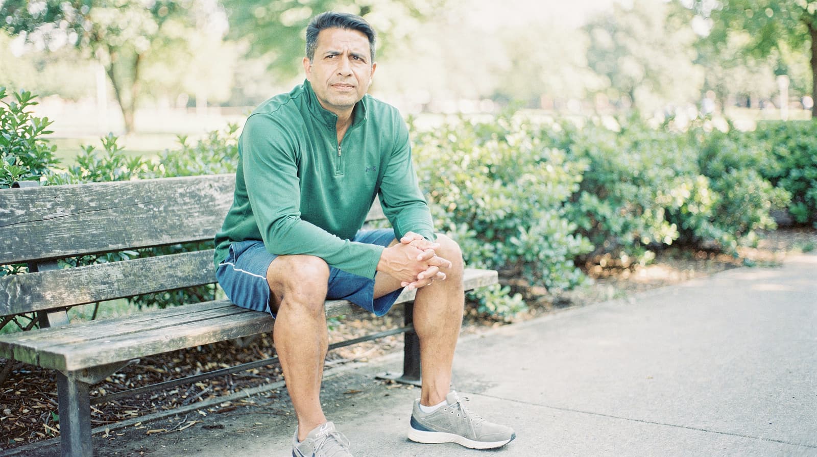 A man in a green shirt and shorts sits on a park bench surrounded by greenery.